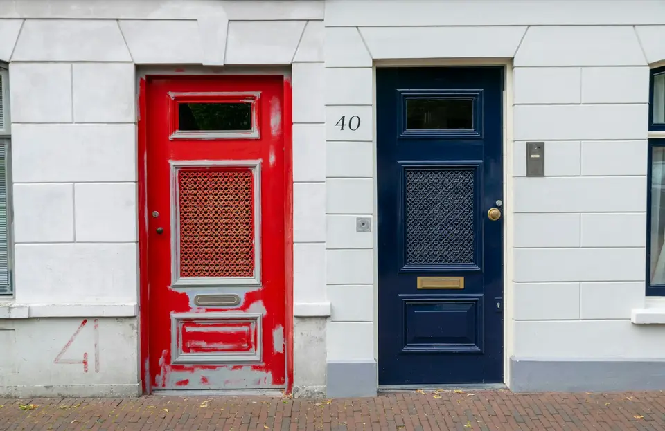 Een rode en een blauwe voordeur met een koperen brievenbus naast elkaar in een witte gevel